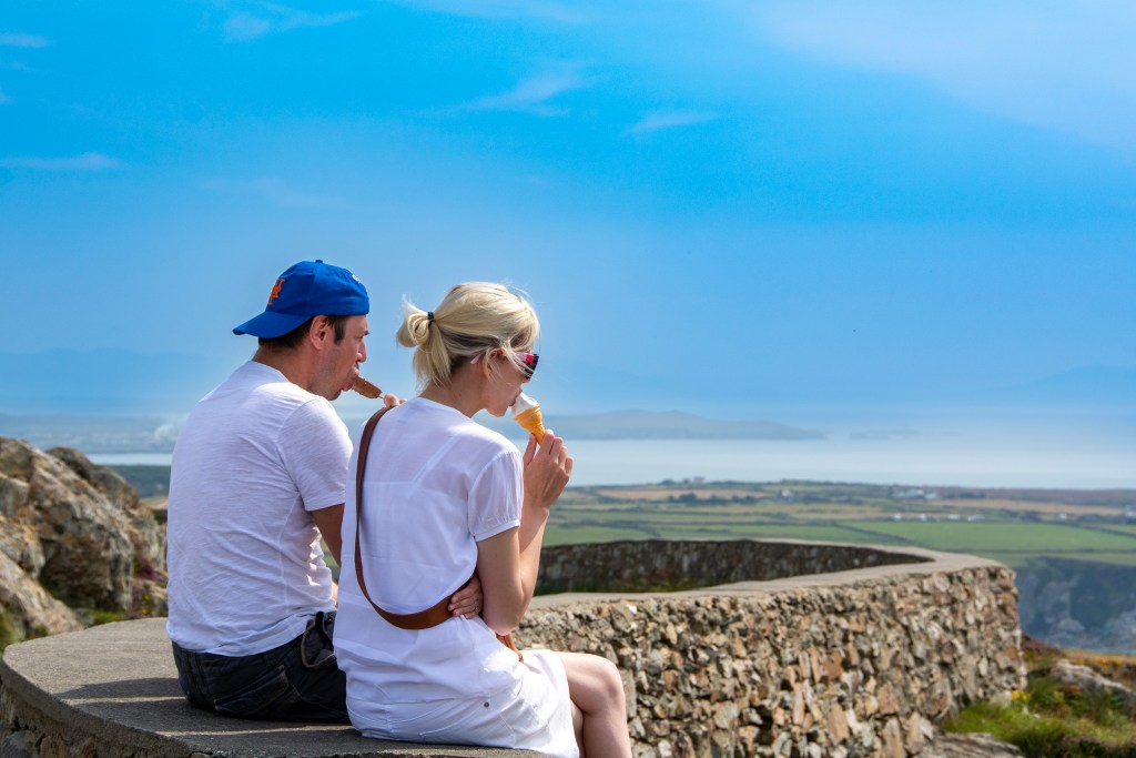 A couple sit looking at the sea eating an ice scream. The sky is bright blue.
